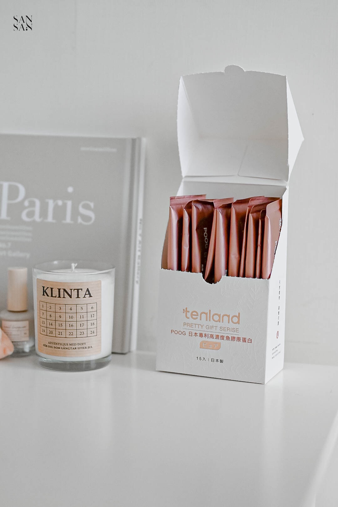 正視角看白色桌上放著一盒打開的膠原蛋白，裡面有數包粉橘色單包裝的膠原蛋白粉，桌上有香氛蠟燭和書