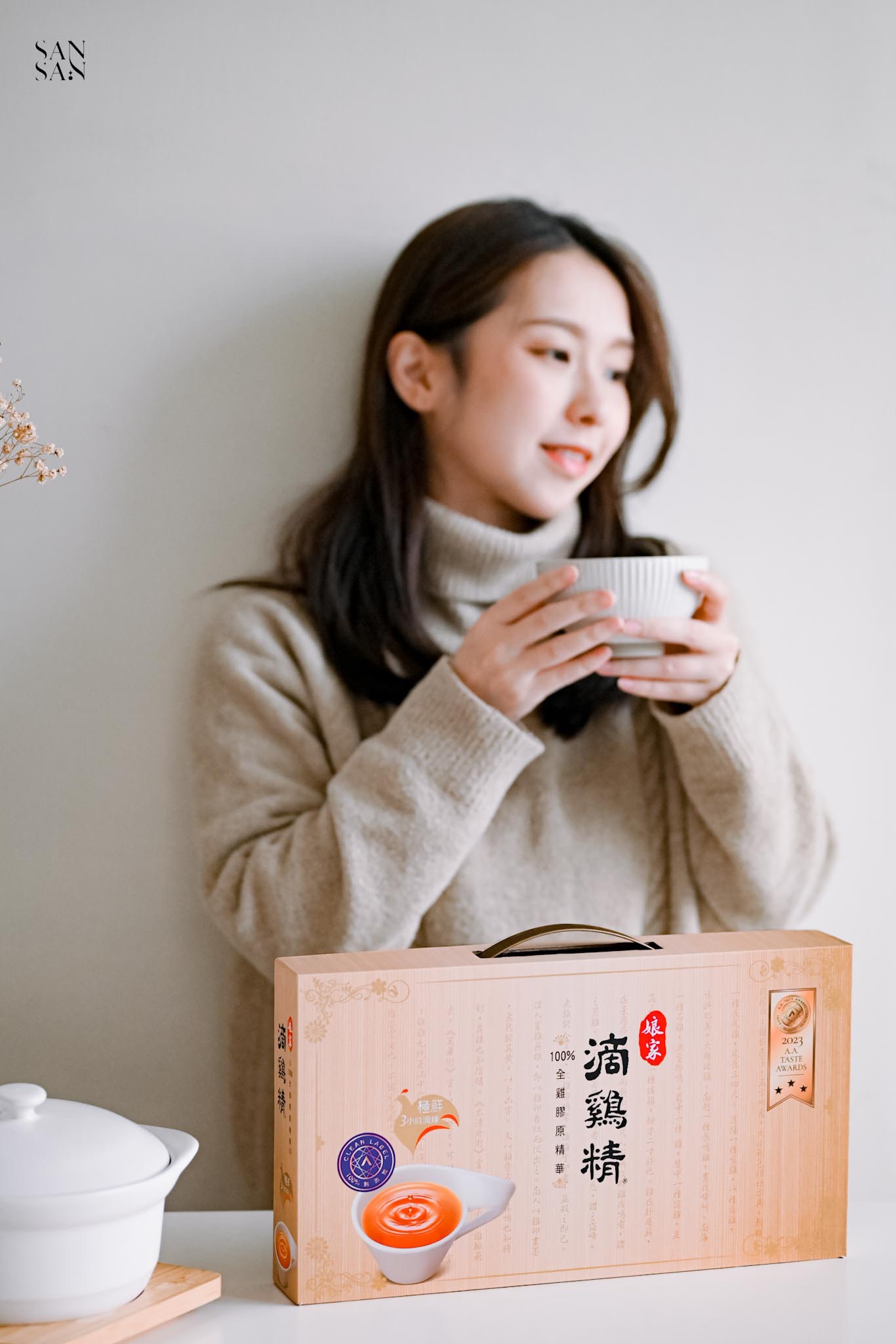 女人微笑端著碗靠在牆上 桌面上擺著娘家滴雞精禮盒