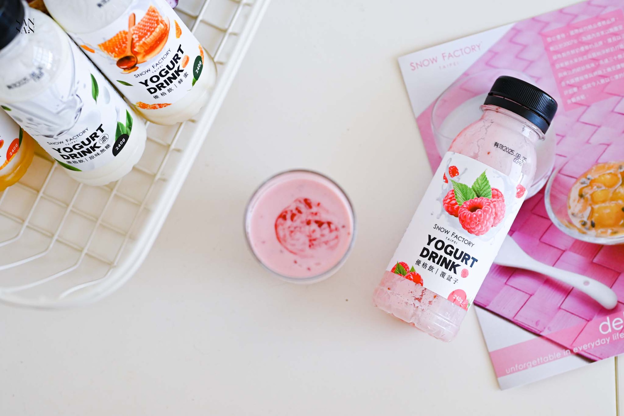 雪坊覆盆子優格飲平放在桌上，旁邊是一杯倒滿粉嫩色優格飲的玻璃杯