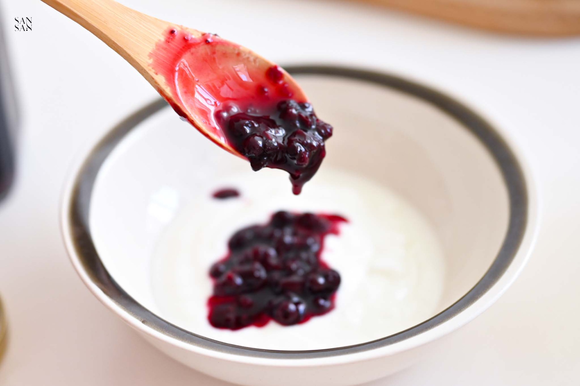 一匙帶有滿滿顆粒的綜合野莓果醬，正在往優格碗裡倒入